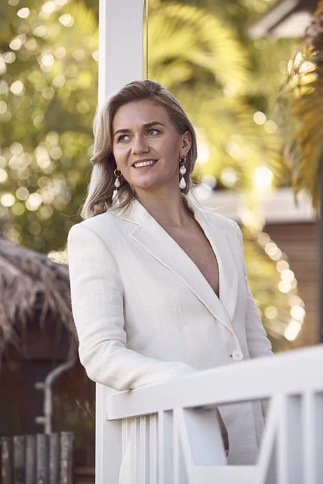 Ariarne Titmus wears a white blazer and leans on the white wooden balcony railing of her family home. Sunlight shines through palm trees behind her.