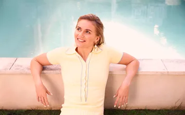 Woman in a light yellow dress sitting by a poolside, smiling and looking upwards.