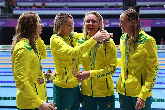 Swimmers Madison Wilson, Kiah Melverton, Ariarne Titmus and Mollie O'Callaghan are standing poolside, dressed in their green and gold uniforms. They hug each other to celebrate setting a world record and winning gold medals.
