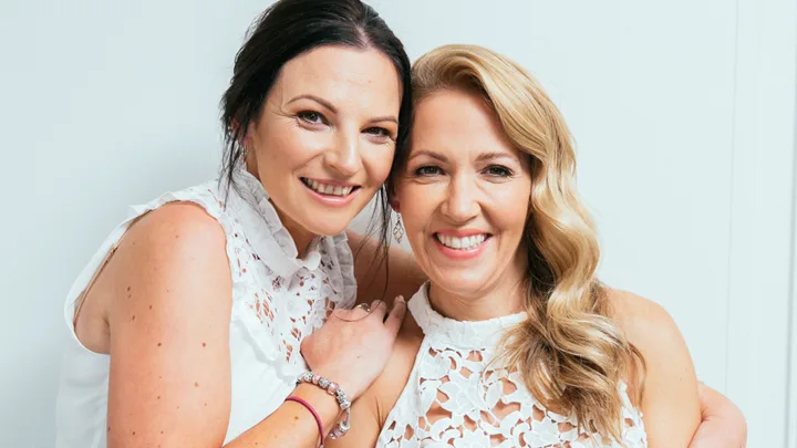 Two women smiling, wearing white lace tops, one with dark hair and the other with blonde wavy hair, posing together.