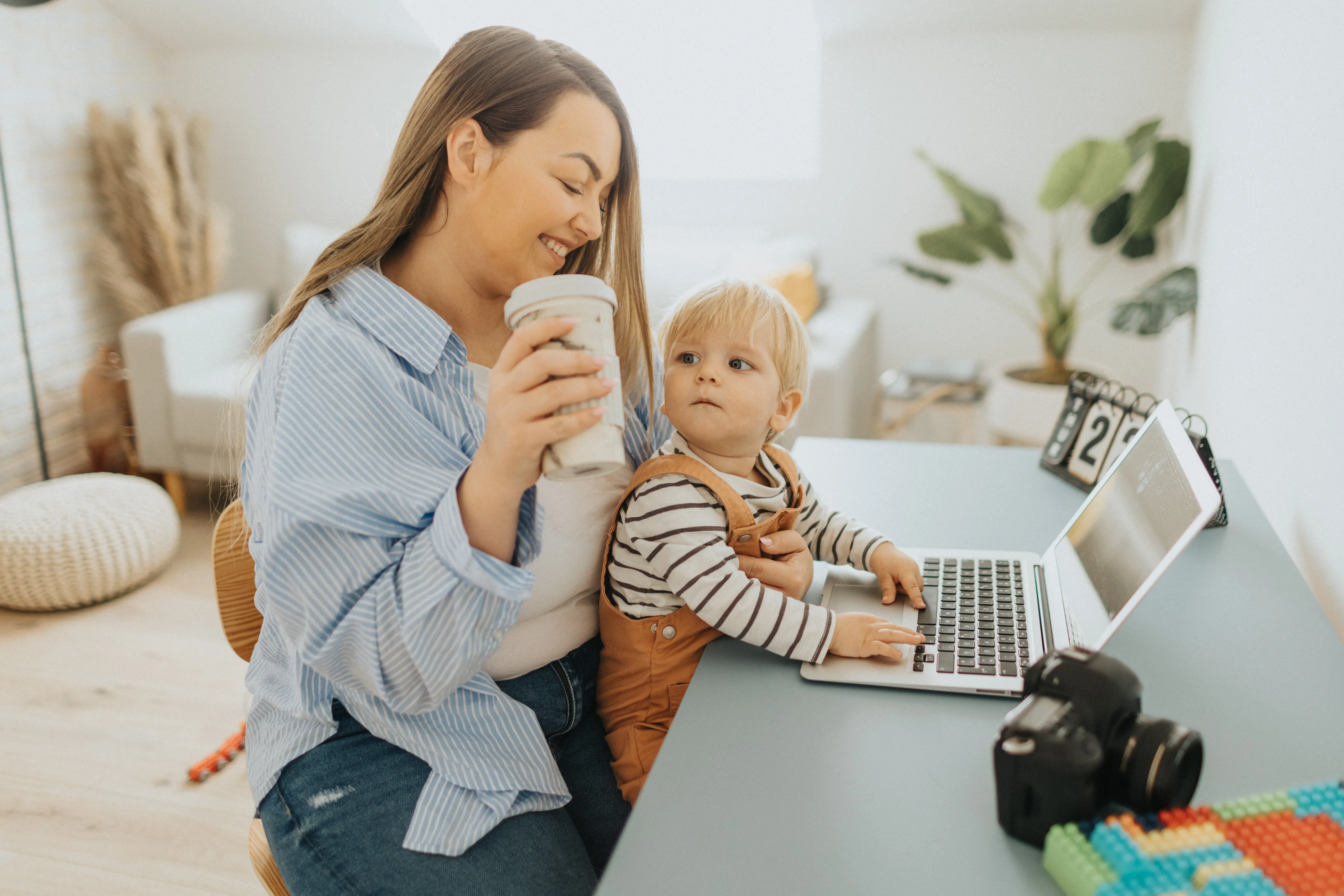 Young mother drinking coffee while working on laptop from home office and holding her baby son on lap