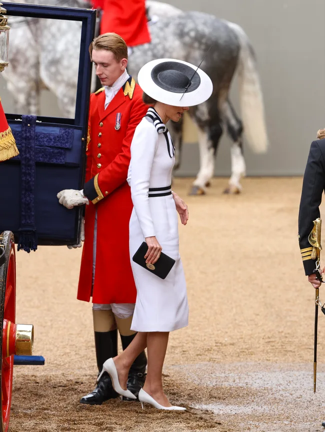 Woman in elegant white dress and hat, stepping out of a carriage assisted by a guard in red uniform at a formal event.