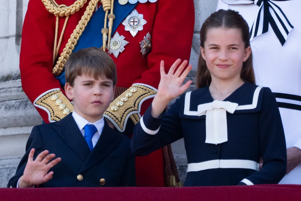 Two young children in formal attire wave from a balcony, with an adult in a decorated red uniform behind them.