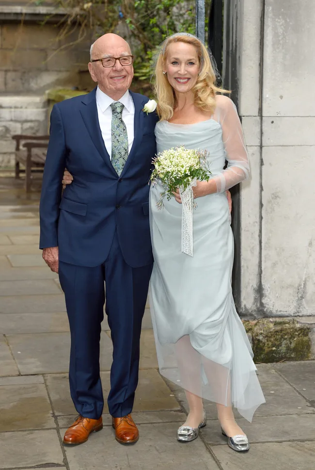 Elderly couple smiling, man in suit, woman in light blue gown holding a bouquet, standing on a stone pavement.
