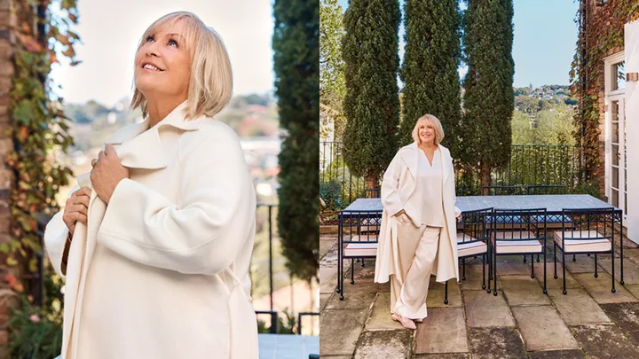 A woman in a cream coat smiles while standing on a patio with trees and outdoor furniture.