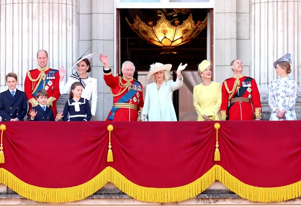 Royal family members wave from a balcony at an official event, wearing formal attire and military uniforms.