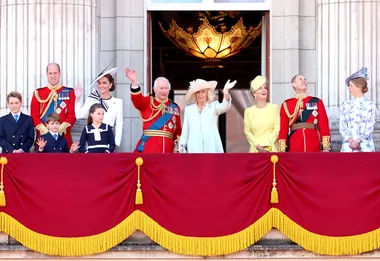 Royal family members wave from a balcony at an official event, wearing formal attire and military uniforms.