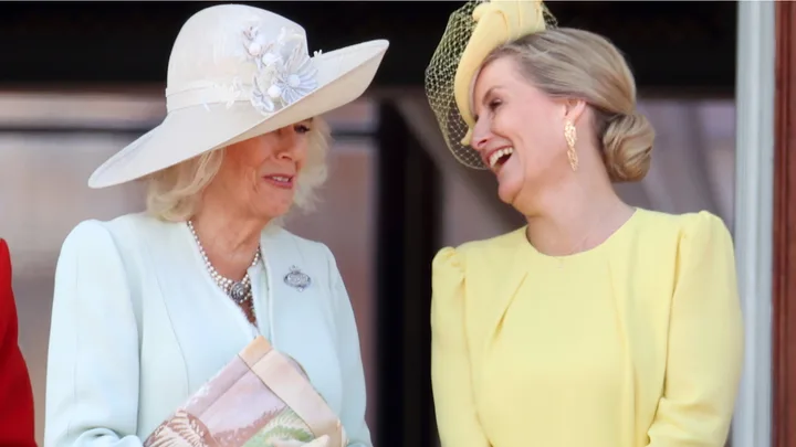 Two women in elegant hats and dresses, smiling and talking to each other outdoors.