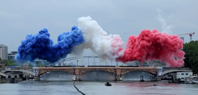 Red, white, and blue smoke over a Parisian bridge, with boats on the Seine River below.