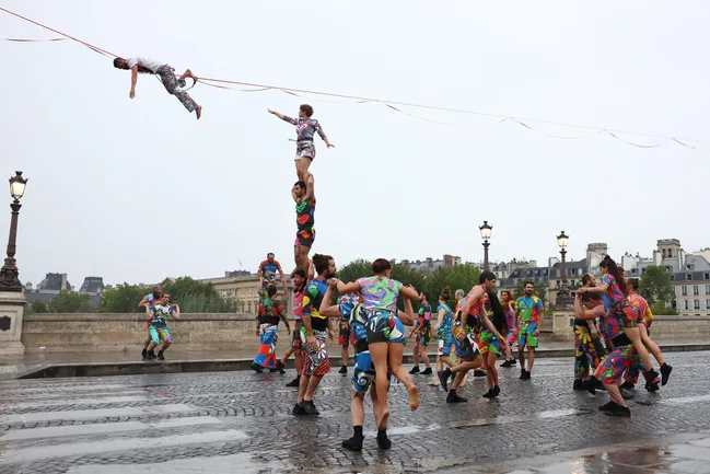Colorful acrobats perform on a cobblestone street with one balanced on a stack of performers, and another on a tightrope.