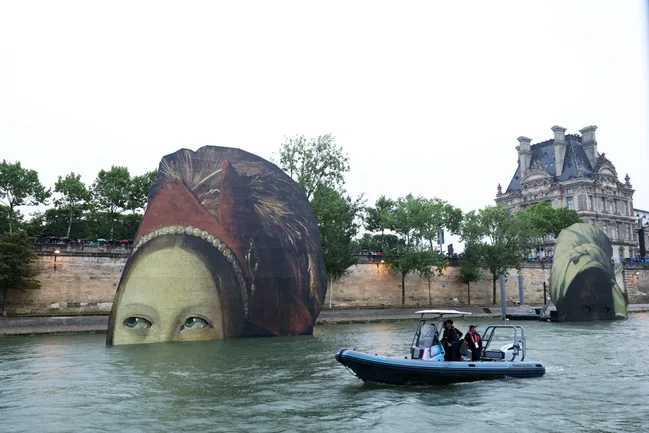 Giant art installations of Renaissance portraits emerge from Seine River in Paris, near the Louvre Museum, with a boat passing by.