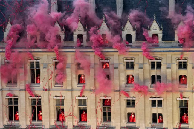 Red smoke fills the air as performers in red cloaks stand at windows of a historic building.
