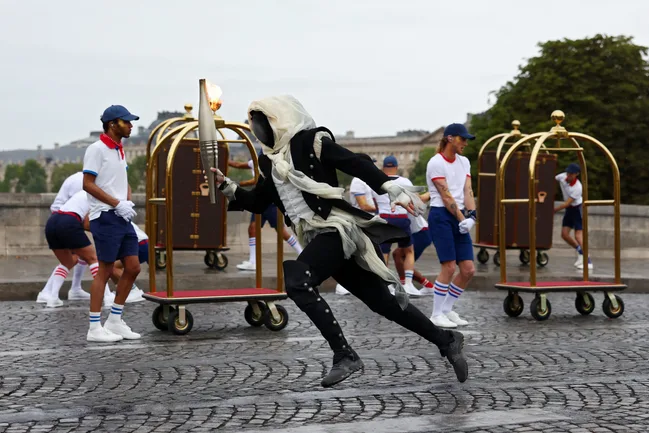 Performer in a dramatic costume sprints with a torch, surrounded by people in themed uniforms and luggage carts.