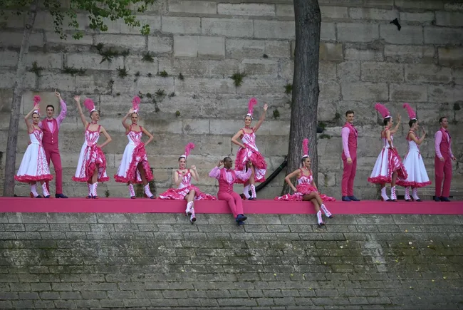 Performers in pink and white costumes pose on a riverside stage against a stone wall.