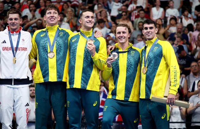 Australian swim team celebrates with medals at the Commonwealth Games, wearing green and yellow tracksuits.