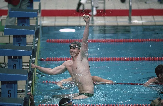 Swimmer jubilantly raises fist in victory at Olympic pool, wearing goggles and swim cap, with lanes visible around him.