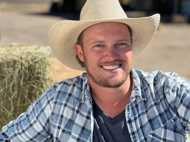 A person wearing a checkered shirt and straw cowboy hat smiles outside with hay bales in the background.
