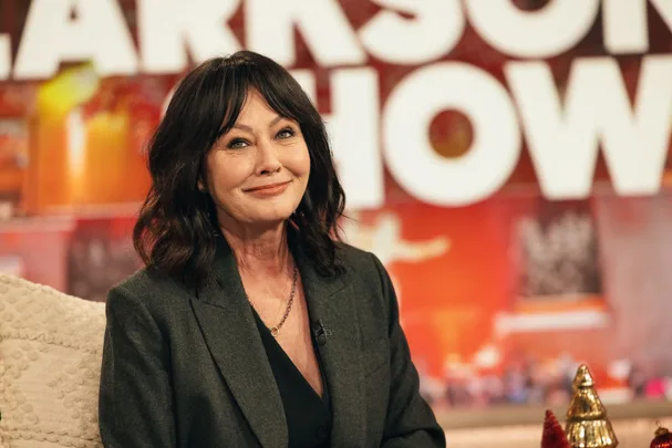 A woman with shoulder-length dark hair smiles, sitting on a talk show set with large blurred letters behind her.