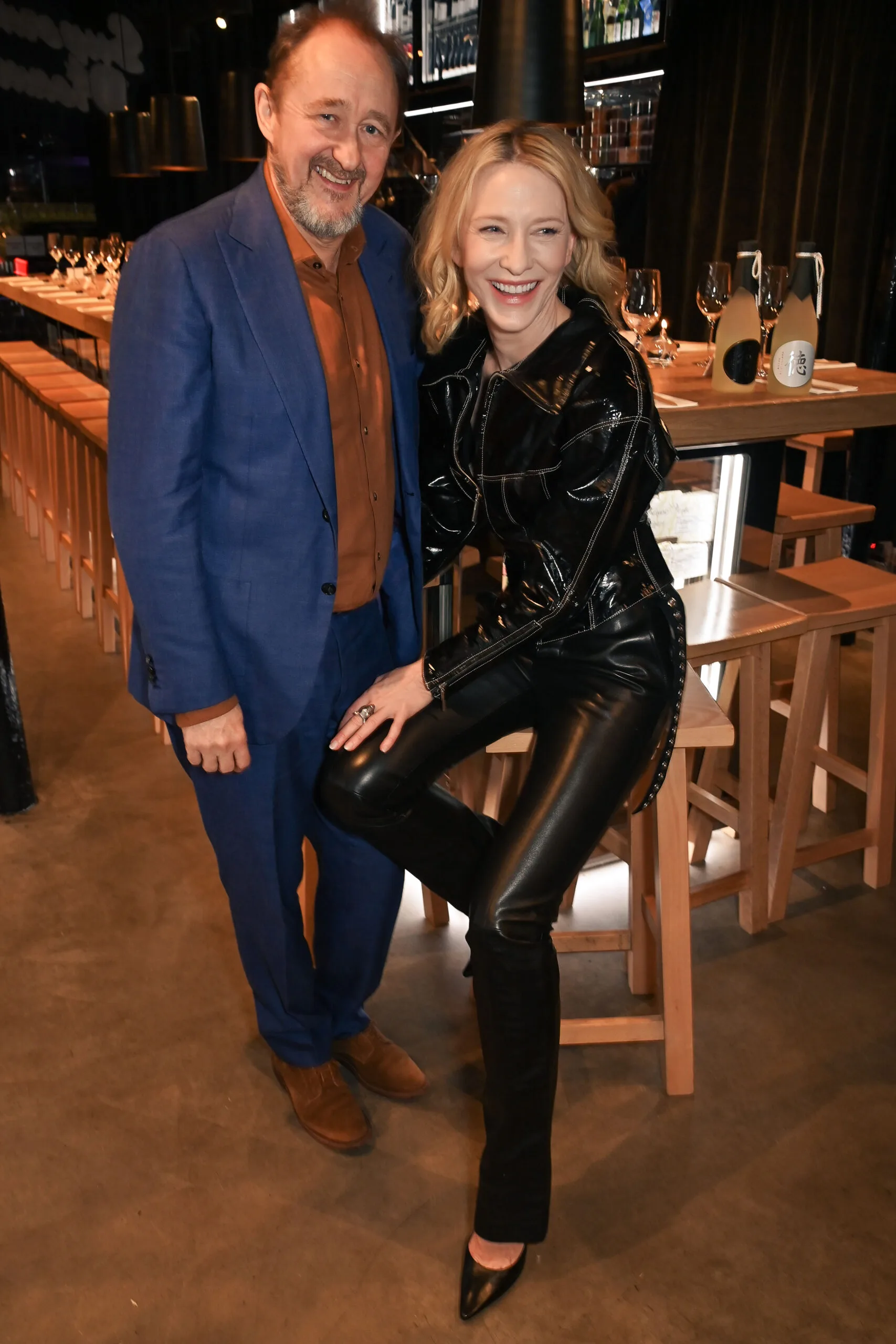 Two people smiling and posing at a stylish bar; one is in a blue suit, and the other in black leather attire.