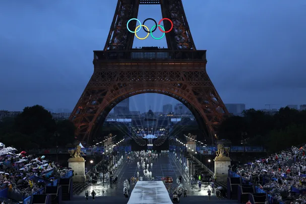 Eiffel Tower at night with Olympic rings, crowds on either side, and a lit-up pathway leading towards Vincennes.