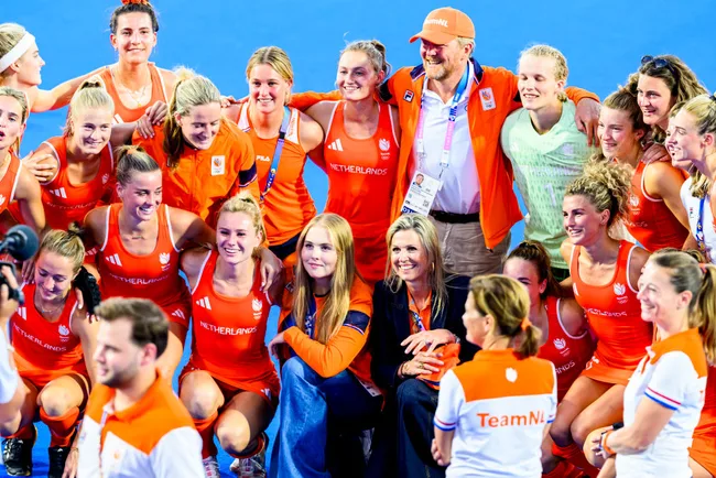 Netherlands women's hockey team smiling in a group photo, wearing orange uniforms on a blue field.