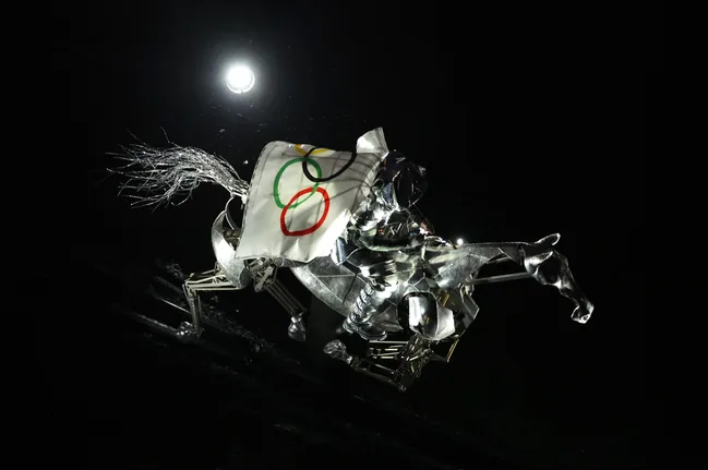 Metallic horse and rider sculpture with Olympic rings flag, illuminated on dark background.
