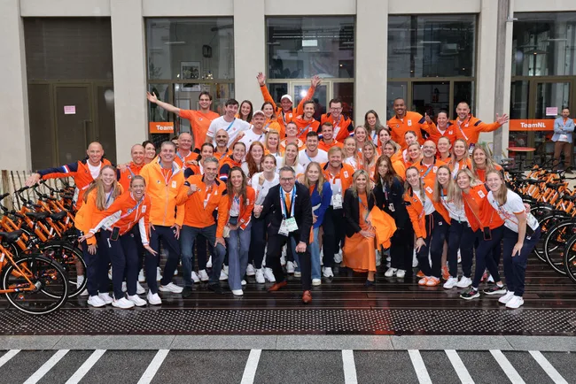 Large group photo of Team NL athletes in orange uniforms, posing enthusiastically outside a building with bicycles.