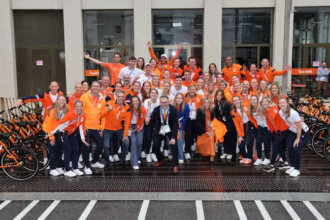 Large group photo of Team NL athletes in orange uniforms, posing enthusiastically outside a building with bicycles.