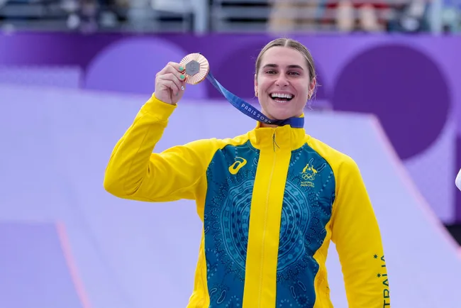 An athlete in an Australian jacket smiles, holding up a silver medal at the Paris 2024 Olympics.