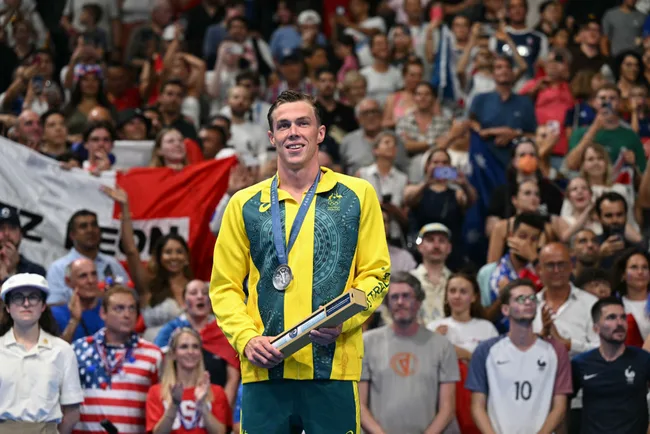 Australian athlete in a green and yellow jacket holds a silver medal, standing in front of a cheering crowd.