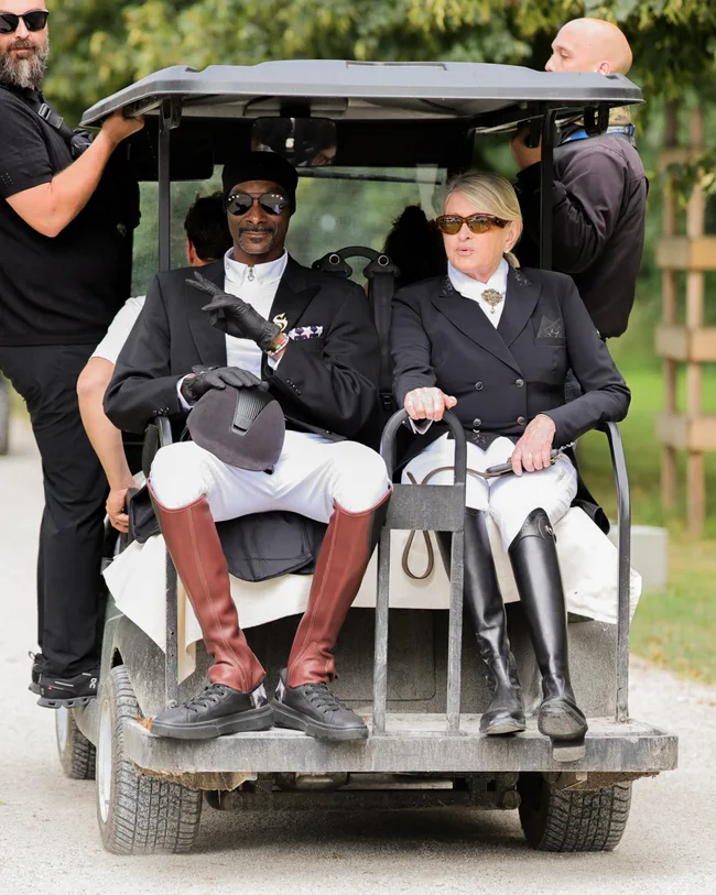 Two people dressed in equestrian attire sitting in the back of a golf cart, with two others standing nearby.