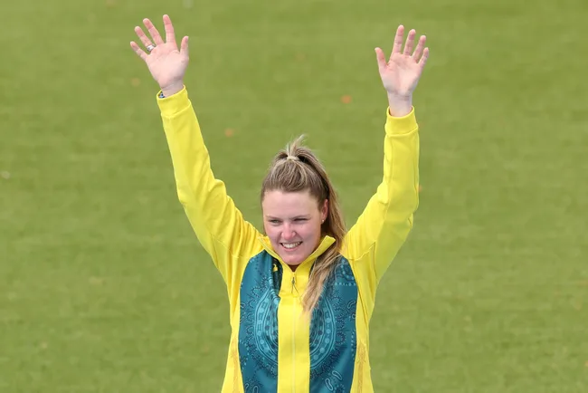 Athlete in a yellow and green jacket raises her arms triumphantly on a sports field.