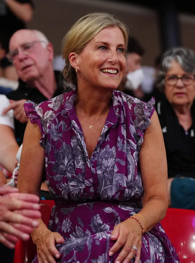 Woman in a purple floral dress smiling at an event; people seated around her.