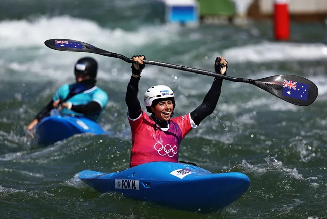 Olympic kayaker celebrates victory holding paddle, wearing Australian flag on kayak, in vibrant blue waters.