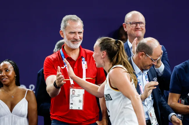 A man in a red shirt claps as a female athlete approaches, surrounded by applauding people against a purple background.