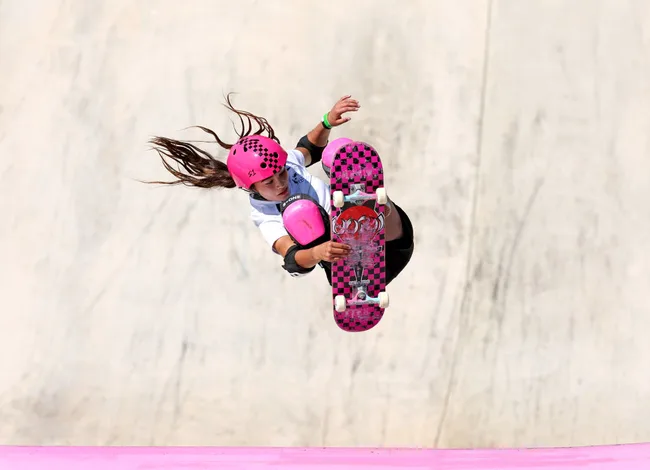 A skateboarder mid-air in a helmet and pads performs a trick on a pink-themed skateboard.