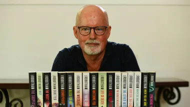 Author with glasses and beard behind a row of books by Michael Robotham, displaying various titles.