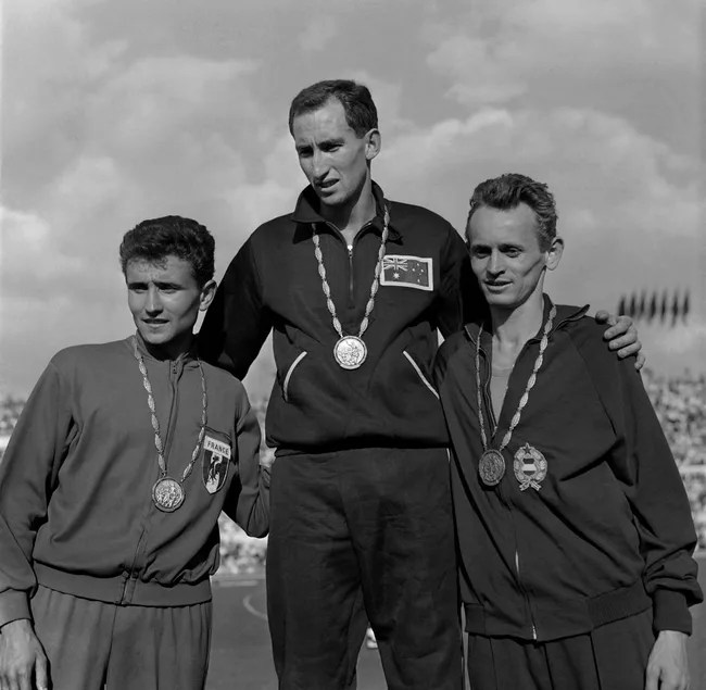 Black and white image of three male athletes on a podium with medals, wearing tracksuits from France, Australia, and Hungary.