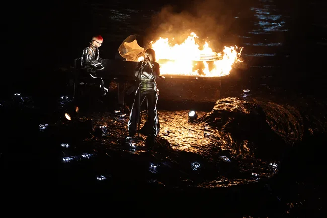 Performers in metallic outfits with a flaming piano on stage during a dramatic live performance.