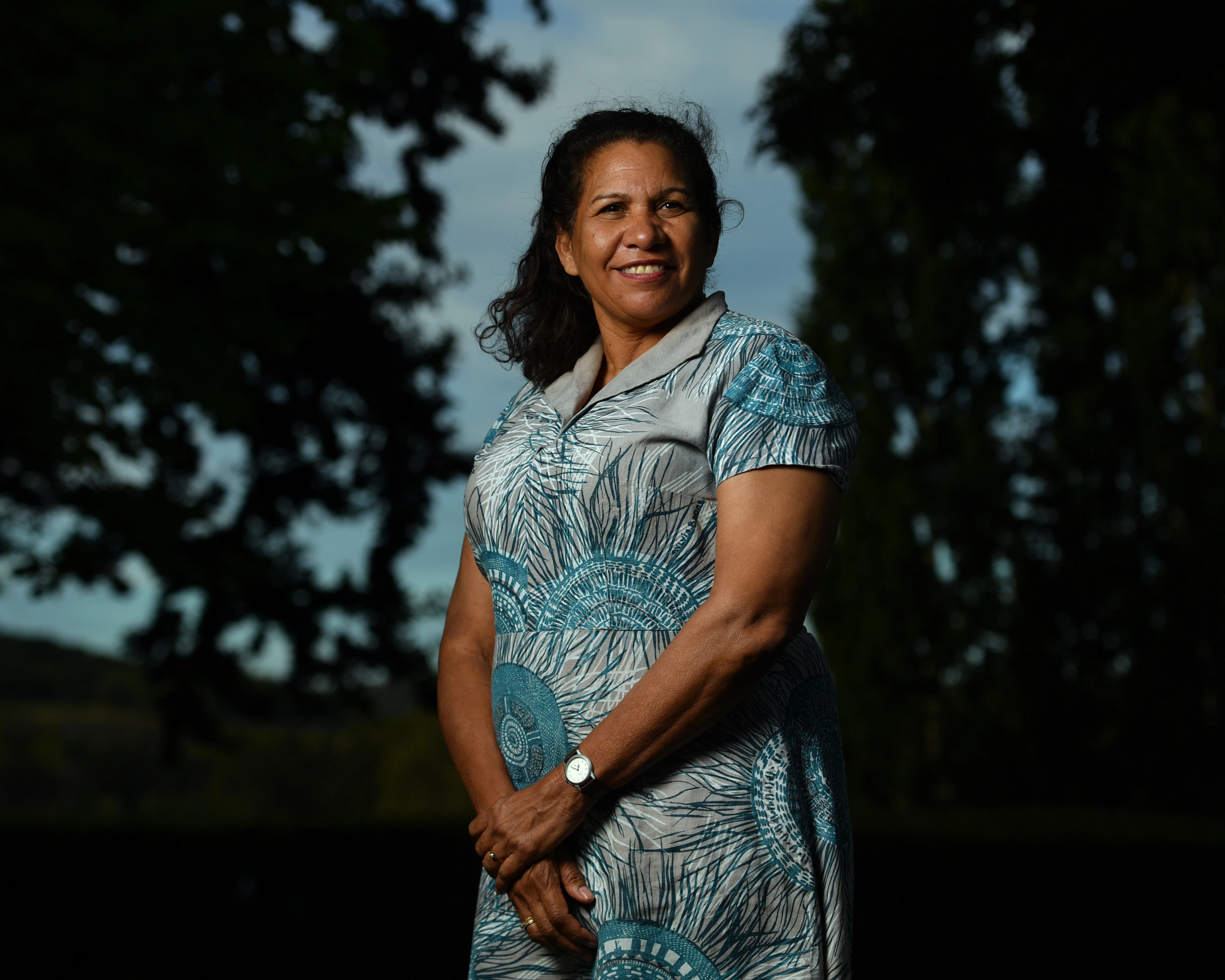 Leanne Liddle stands smiling thoughtfully in front of a row of trees and a pale blue sky. She wears a dress with a blue and grey print. Her hair is dark brown and wavy and hangs just below her shoulders.