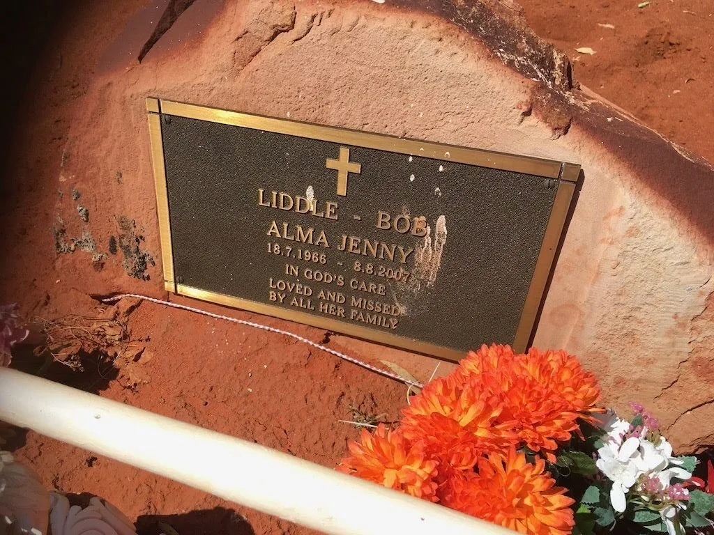 The gravestone of Jenny Alma Liddle-Bob sits against a rock. Brightly coloured flowers have been laid beside it.