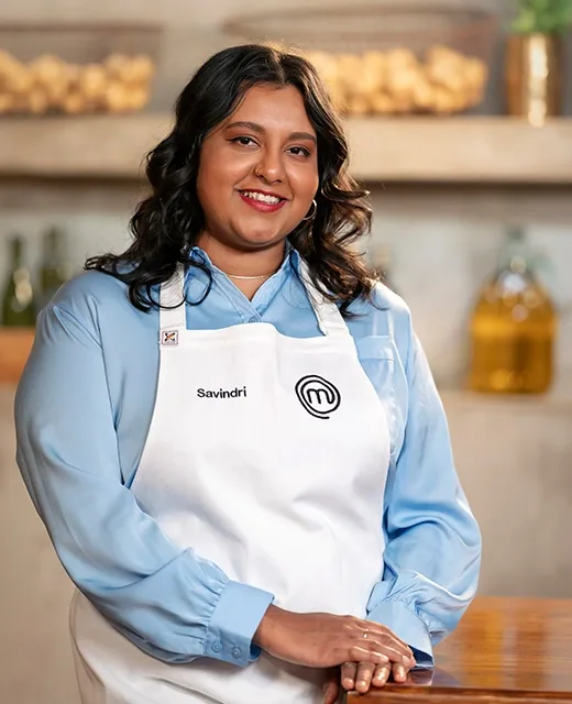 Savindri, a contestant in a MasterChef apron, smiles standing in a kitchen setting.