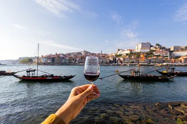 Hand holding a glass of red wine overlooking Douro River with boats and buildings in Porto, Portugal.