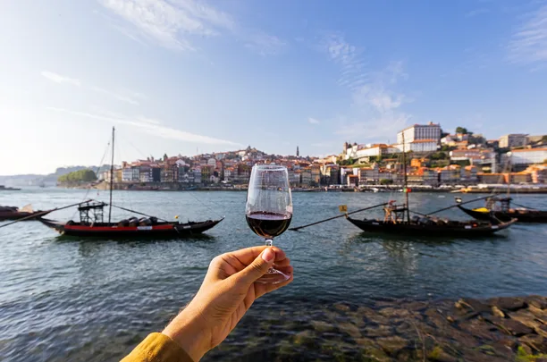 Hand holding a glass of red wine overlooking Douro River with boats and buildings in Porto, Portugal.