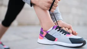 Person tying the shoelaces of a colorful sneaker, wearing black leggings outdoors.