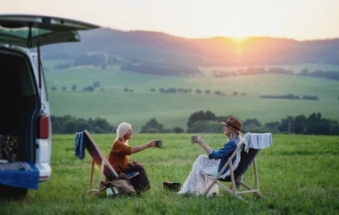 Two people sitting in chairs, enjoying drinks near a van in a scenic field at sunset.