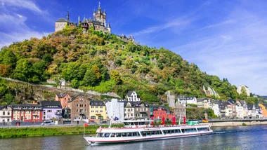 Riverboat cruising near Cochem Castle, surrounded by colorful houses and lush greenery under a clear blue sky.