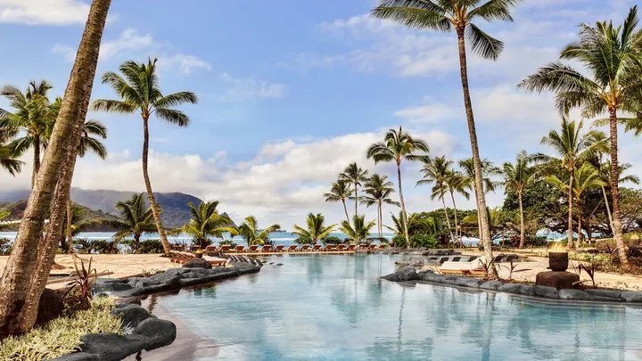 Tropical resort pool with palm trees and mountains in the background under a bright blue sky.