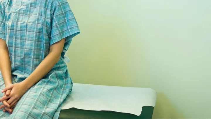 Person in a blue hospital gown sits on an exam table against a plain wall.