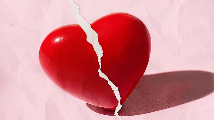 A red heart with a jagged white paper tear running vertically, set against a pink textured background.